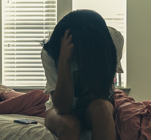 Depressione, tipi e come si manifesta. Donna seduta sul letto con la fronte rivolta verso il basso e le mani sulla testa