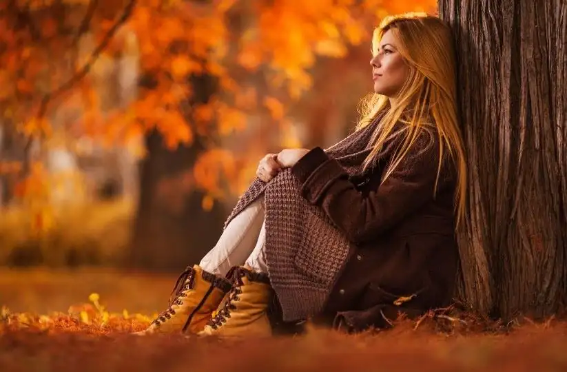 Solitudine interiore, giovane donna sola seduta con la schiena appoggiata ad un albero nel mezzo di un paesaggio autunnale che suscita malinconia